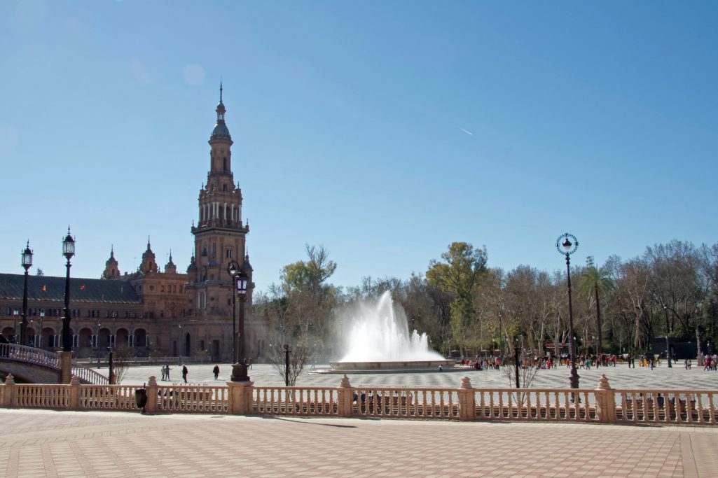 Fontaine au centre d'une place, avec un bâtiment historique et un ciel bleu dégagé.