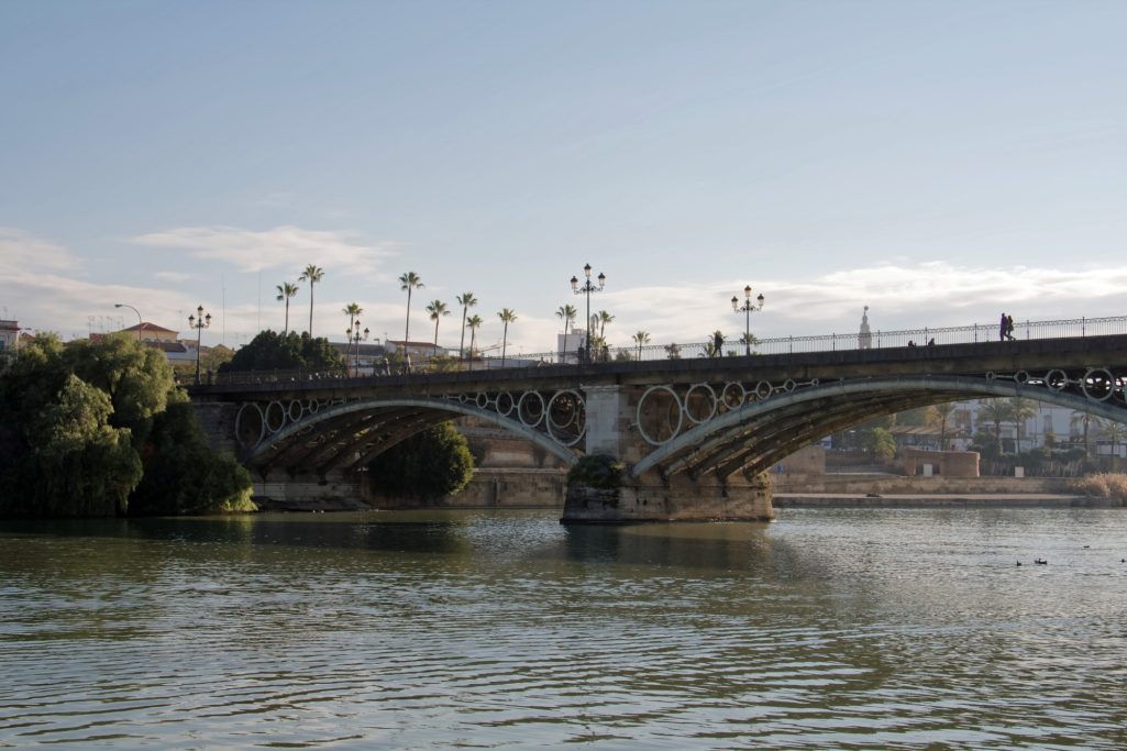 Pont en arc sur une rivière, avec des palmiers en arrière-plan et des passants.